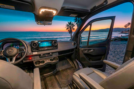 Interior of an RV rental van with its door open, offering a serene view of a vibrant beach sunset over the ocean and palm trees.