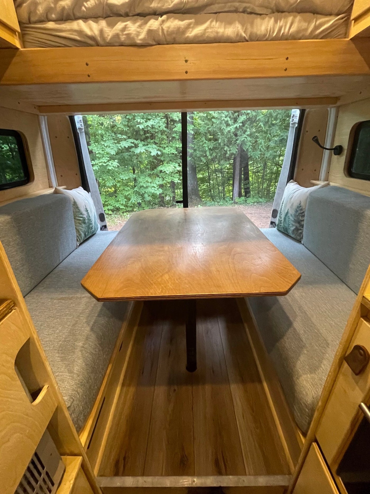 Inside a cozy RV rental: a wooden dinette with a forest view, comfy benches, and an overhead bed. — photo 7