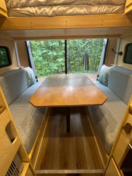 Inside a cozy RV rental: a wooden dinette with a forest view, comfy benches, and an overhead bed.