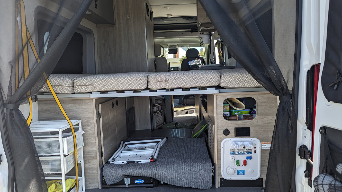 Spacious RV rental interior from the rear, showing a raised bed, storage, and utility panel for compact camper van living.