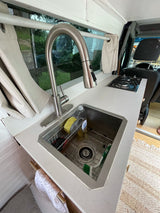 Compact RV rental kitchen features a sink, cooktop, and white counter in a functional van conversion setup.
