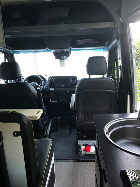 Inside an RV rental: view from rear showing driver's cockpit, seating, and a compact kitchen area with sink & fire extinguisher.