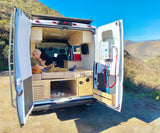 RV rental van life: Man works on laptop in custom camper, doors open to mountains & nature. Includes water heater.