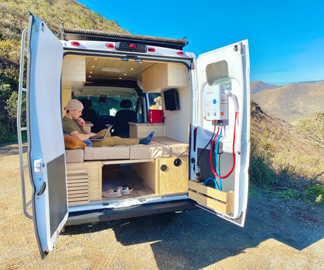 RV rental van life: Man works on laptop in custom camper, doors open to mountains & nature. Includes water heater.