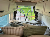 Relax in a cozy RV rental: This image shows a comfortable bed inside an RV, looking out the back to a park-like view with trees and a paved path.