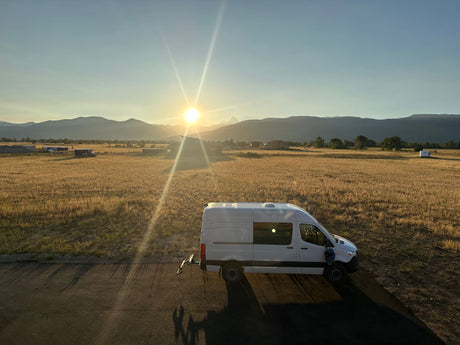 White RV rental van at sunset, parked beside a golden field with distant mountains. Perfect for adventure.