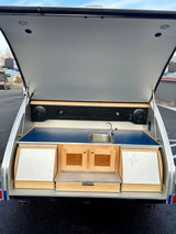 This RV rental teardrop trailer shows its open rear hatch revealing a full outdoor kitchen with sink, cabinets, and speakers.