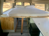 Interior of an rv rental camper van, featuring a cozy bed with a white mattress and pillows, built-in storage, and a portable fridge.