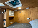 Cozy RV rental interior: wooden walls, bed, window AC, ceiling fan, and panel controls for your travel comfort.