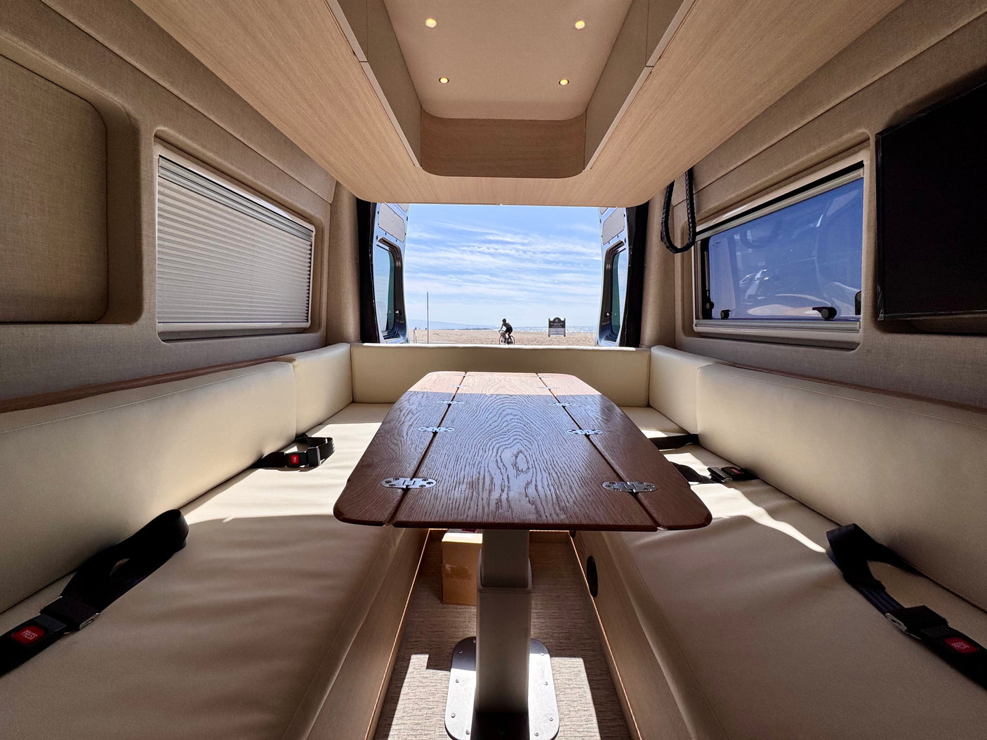 Inside an RV rental: a wooden table & comfy seats with a stunning view of a sunny beach & bicyclist through the open back.