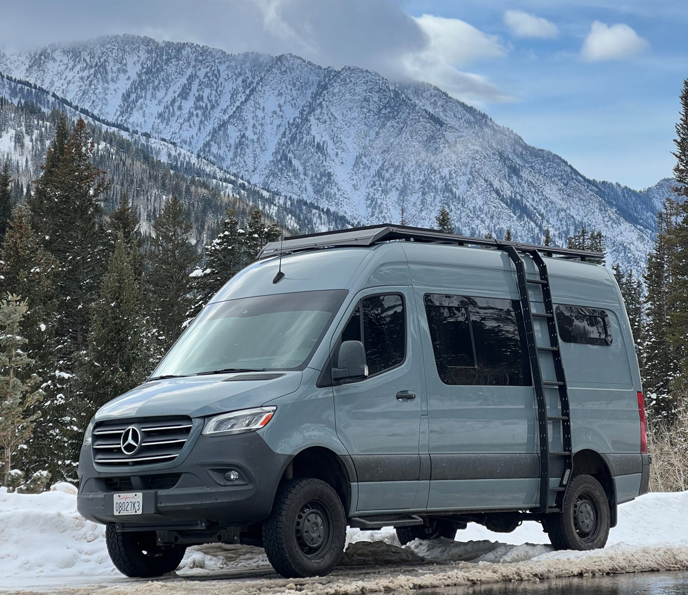 Sleek grey RV rental: a Mercedes Sprinter van parked on a snow-covered road with majestic, pine-dotted mountains. Perfect for winter adventures.