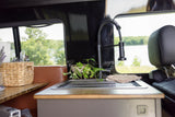 RV rental kitchen area features a sink, modern faucet, plants, and utensils, with a scenic lake and tree view outside.