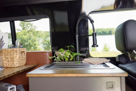 RV rental kitchen area features a sink, modern faucet, plants, and utensils, with a scenic lake and tree view outside.
