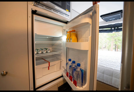 RV rental interior: Open fridge with drinks (OJ, soda, water), microwave above. A cozy bed is visible, ready for your adventures.