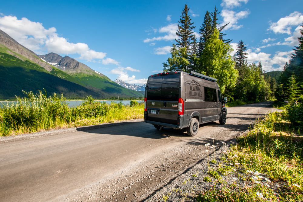 Experience nature with an RV rental! A dark camper van is parked on a dirt road by a serene lake & majestic snow-capped mountains.