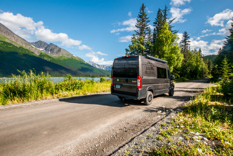 Experience nature with an RV rental! A dark camper van is parked on a dirt road by a serene lake & majestic snow-capped mountains.