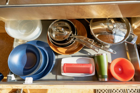 Inside an RV rental drawer, neatly organized with stacked pots, pans, blue dishes, plastic bowls, a red speaker, and green tumbler.