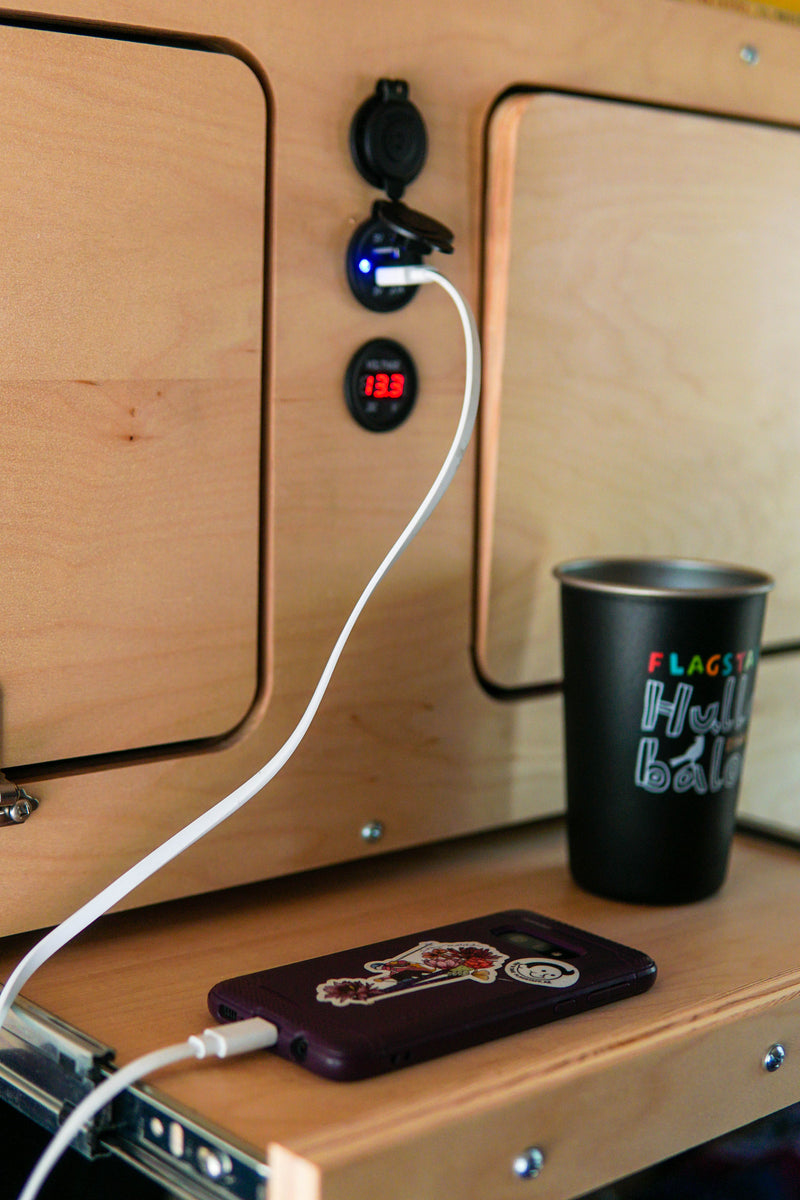 RV rental interior: phone charging from a built-in USB port, next to a cup on a wooden surface. System voltage: 13.3V. — photo 11