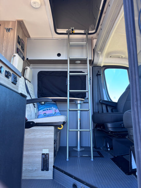 Interior of an RV rental: a ladder to the pop-top bed, textured floor, comfortable seating, and visible control panels.
