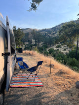 An rv rental set up for camping: white camper van, blue chairs, and a colorful mat overlook a dry grassy hillside.