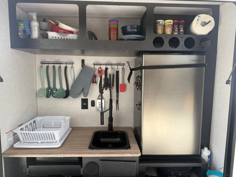 Organized RV rental galley kitchen with a compact fridge, sink, hanging utensils, and upper shelf storage.