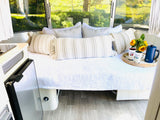 Relaxing RV rental interior: A comfy bed with decorative pillows & sunflowers, brightened by nature outside the large windows.