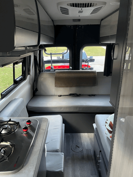 Inside this modern RV rental: a compact kitchen with stove, lounge seating, overhead storage, and AC for comfort.