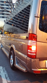 Tan RV rental van, rear view with an illuminated taillight, reflecting urban architecture and trees on its shiny surface.