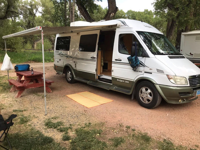 Relaxing at a dirt campsite with an RV rental, featuring an extended awning, picnic table, and outdoor mat.