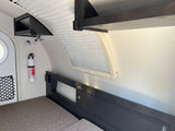 Compact RV rental interior: bed, wall-mounted storage, window with blinds, and fire extinguisher.