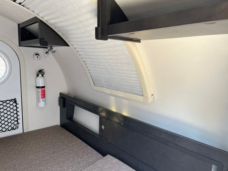 Compact RV rental interior: bed, wall-mounted storage, window with blinds, and fire extinguisher.