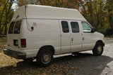 A white high-top Ford Econoline camper van, suitable for an rv rental, parked on a gravel path with autumn leaves.