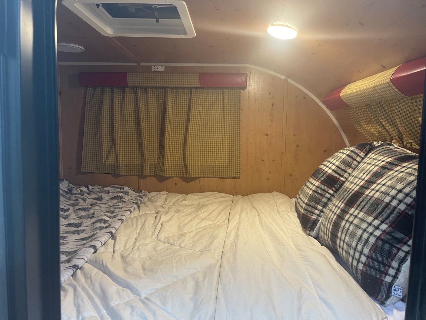 Cozy RV rental interior featuring a comfortable bed with plaid pillows, wooden walls, and a window with checkered curtains.