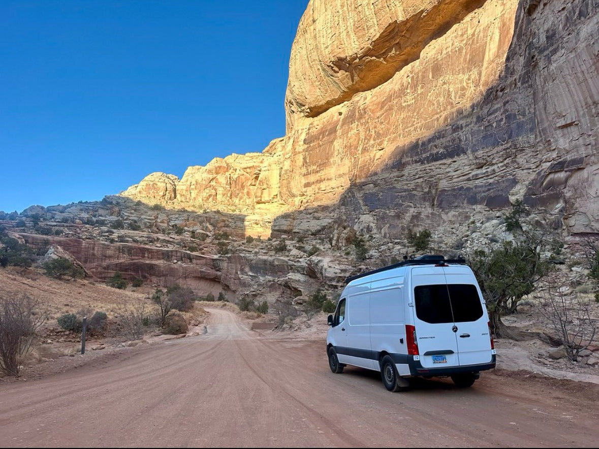 White camper van on a dirt road by towering red rock cliffs, an RV rental exploring a desert park.