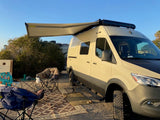 Perfect day at a campsite! This rv rental, a cream-colored camper van with its awning out, offers comfortable outdoor living.