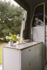 An RV rental's modern interior kitchen with a sink, faucet, potted plant, and control panel, offering a view of lush trees.