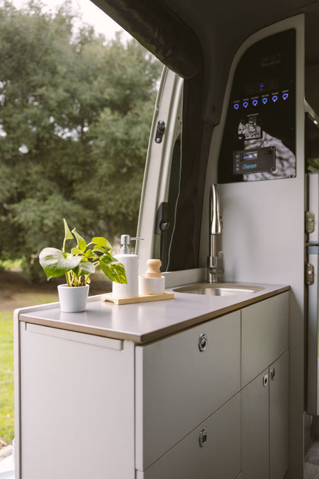 An RV rental's modern interior kitchen with a sink, faucet, potted plant, and control panel, offering a view of lush trees.