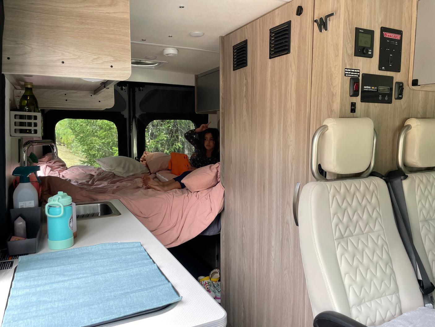 Cozy RV rental interior shows a bed with pink sheets, kitchenette, passenger seats, and a child relaxing.