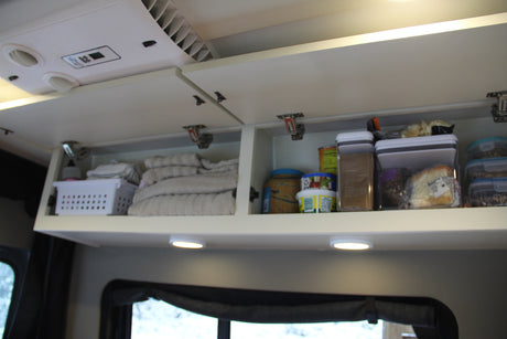 Organized RV rental overhead storage with folded towels, food containers & a basket. Ceiling AC unit visible.