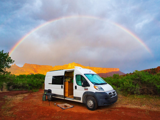 Vivid rainbow over a white campervan RV rental. Parked on red dirt near sunlit red cliffs, ideal for scenic travel.