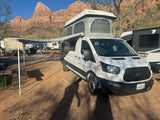 White camper van (an RV rental) with a pop-top, parked at a sunny campground against stunning red rock mountains.