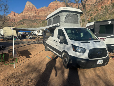 White camper van (an RV rental) with a pop-top, parked at a sunny campground against stunning red rock mountains.
