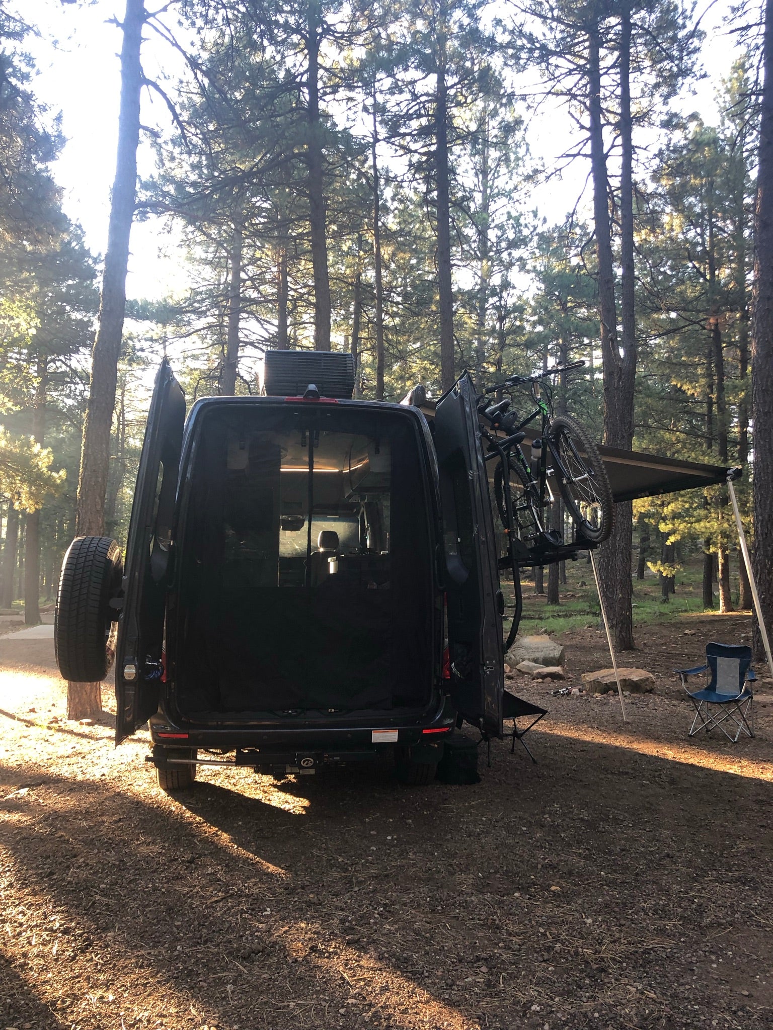 RV rental van campsite in a sunny forest. Open doors reveal gear, bikes ready for trails, and an awning provides shade.