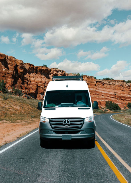 Front view of a white Mercedes Sprinter RV rental van on a desert road with grand red rock cliffs under a cloudy sky.