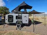 This RV rental shows a gray camper with an open rooftop tent, awning, and chairs set up by a scenic lake.