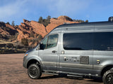 This grey RV rental van, equipped for adventure with large tires, is parked by red rock formations under a blue sky.
