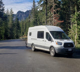White Ford Transit van, ideal for an RV rental, parked on a road amid towering trees and mountains. Van life journey.