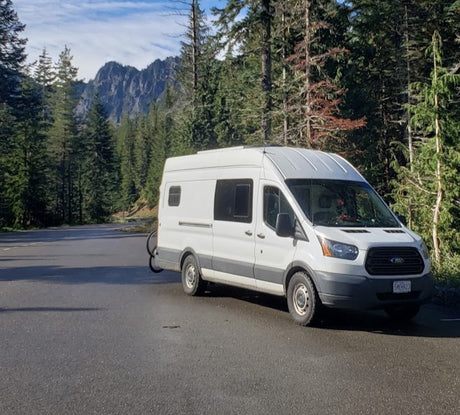 White Ford Transit van, ideal for an RV rental, parked on a road amid towering trees and mountains. Van life journey.