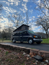 A dark blue pop-top Ford van, like an RV rental, parked on asphalt with bare trees under a cloudy sky.