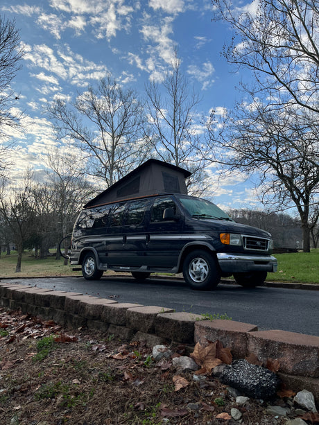 A dark blue pop-top Ford van, like an RV rental, parked on asphalt with bare trees under a cloudy sky.